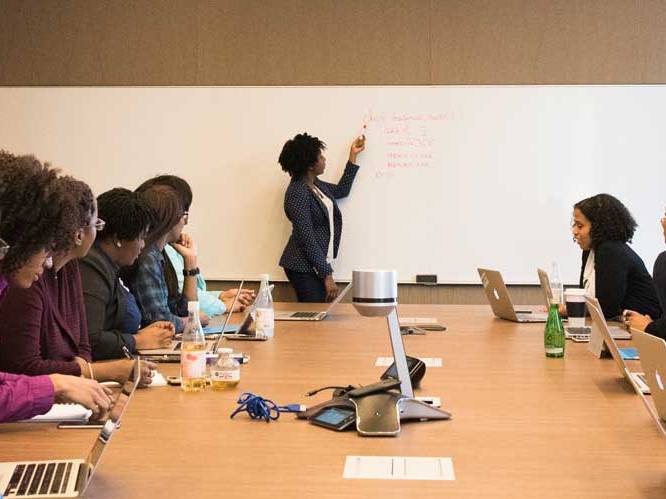 Woman is pointing to the white board, talking to the group of students who are focusing on her