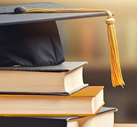 A KSU graduation cap resting on top of a stack of books that are black with a gold book in the middle.