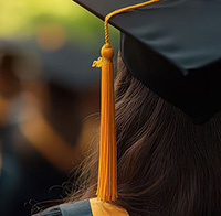 Close-up of a KSU grad student with graduation cap on a student, focusing on the shiny golden tassel and the year charm, with blurred fellow graduates in the background at an outdoor ceremony.