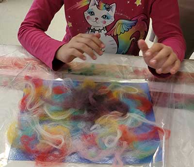  / Still photograph of a student layering colorful wool for a wet felted vessel.
