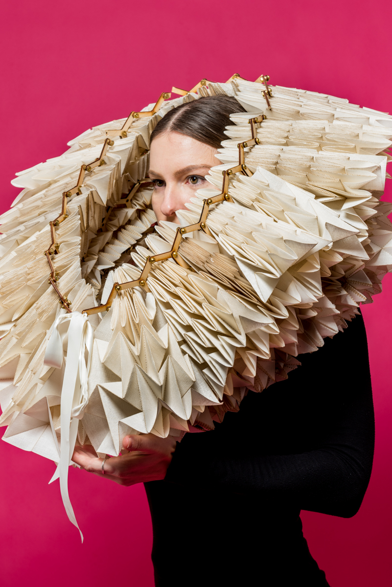 A photograph of a woman holding an object on her head in front of a pink backdrop