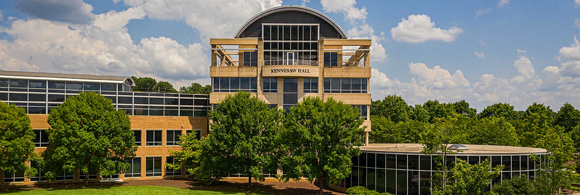 Kennesaw Hall at Kennesaw State University on a sunny day. The building has a curved roof and large windows, surrounded by trees and a grassy area.