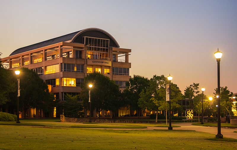 A photo of Kennesaw Hall at Kennesaw State University at dusk. The building is illuminated with warm lights, and there are trees and a grassy area in the foreground.