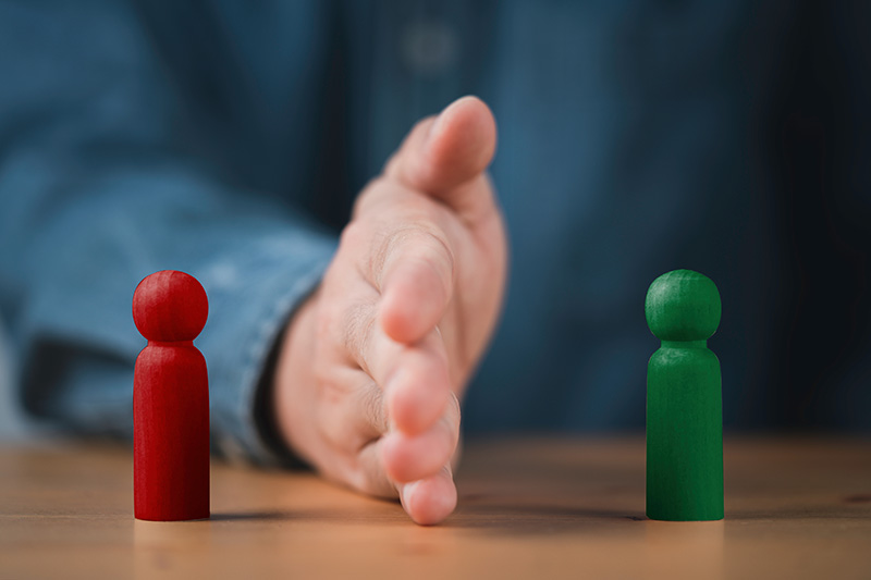 A hand reaching between two wooden figures, one red and one green, on a wooden table representing a conflict.