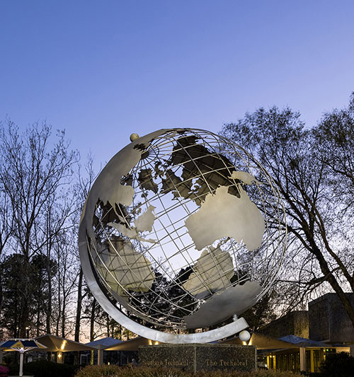 Marietta KSU campus globe is a large metallic globe sculpture set against a blue sky with trees and a buildings in the background.