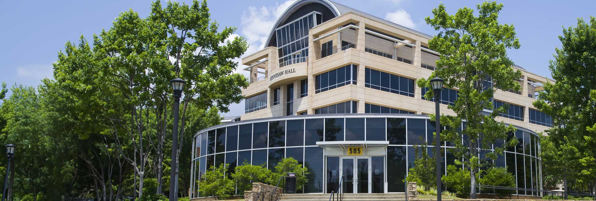 Kennesaw State University's, Kennesaw Hall, a modern building with a curved glass facade and a large arched window. Trees line the sides of the building, and a sidewalk leads up to the entrance.