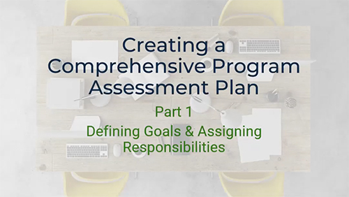 A top-down view of a conference table with a laptop, keyboard, mouse, pens, and papers scattered across it. The text "Creating a Comprehensive Program Assessment Plan" and "Part 1 - Defining Goals & Assigning Responsibilities" is written on the table. Two yellow chairs are visible on either side of the table.