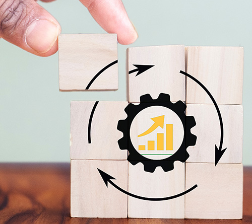 A hand placing a wooden block on a stack of blocks, forming a puzzle with a gear and an upward-facing arrow symbolizing growth and process improvement.