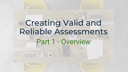 A top-down view of a conference table with a laptop, keyboard, mouse, pens, and papers scattered across it. The text "Creating Valid and Reliable Assessments" and "Part 1 - Overview" is written on the table. Two yellow chairs are visible on either side of the table.