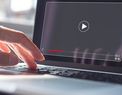 A hand hovering over a laptop keyboard, with a video player interface displaying a play button and a red progress bar.