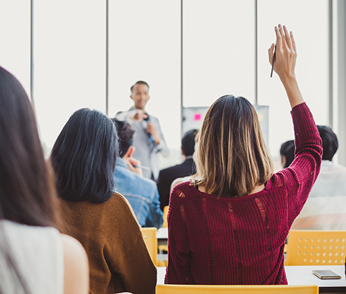 A small classroom of people in a workshop and training class with a woman with her hand raised and the instructor calling on her.