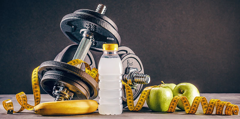 A still life image of fitness equipment and healthy food, including dumbbells, a measuring tape, a water bottle, a banana, and apples