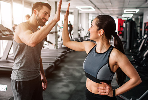 A trainer and a trainee high-fiving in a fitness center. Both are wearing workout attire, and they appear cheerful and motivated. Treadmills and other gym equipment are visible in the background.