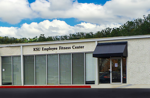 Exterior view of the KSU Employee Fitness Center building, featuring large windows, a black awning over the entrance, and a sign reading "KSU Employee Fitness Center" on the facade.