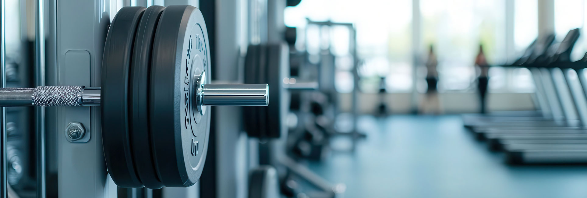 A close-up of a barbell with weight plates in a modern gym. In the background, treadmills and other exercise equipment can be seen.