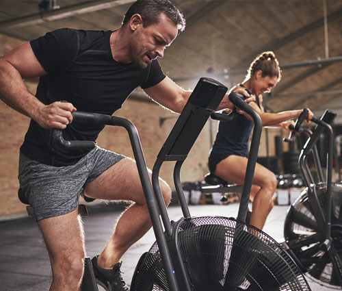 A man and a woman intensely working out on stationary air bikes in a gym, focusing on building strength and stamina. 