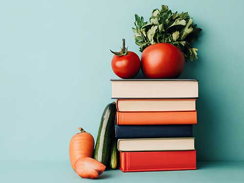 A stack of books with a tomato and parsley on top and a carrot and cucumber in front. The background is a light blue.