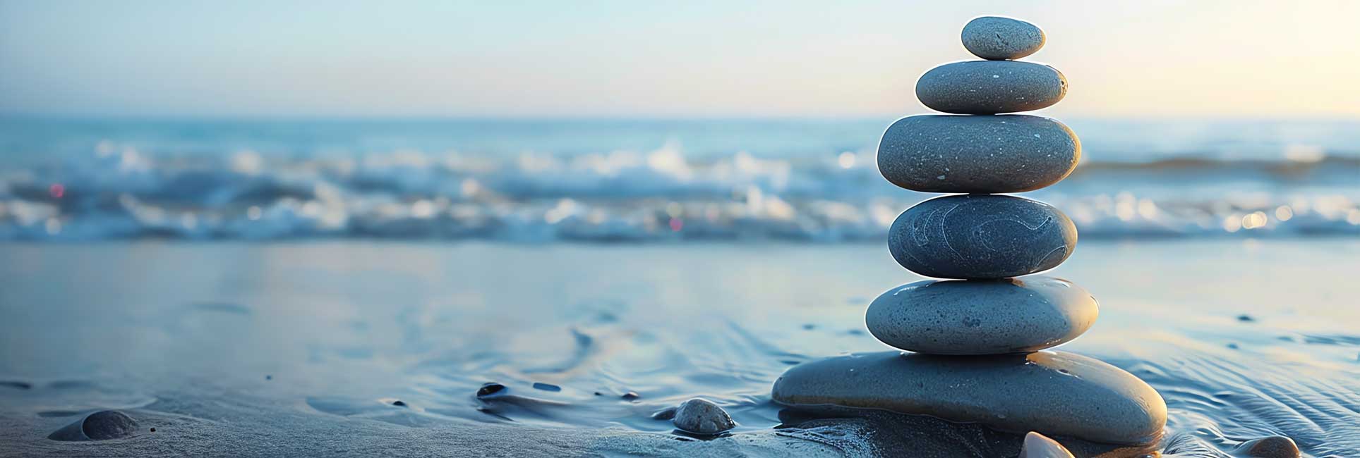 A stack of six gray stones balanced on a sandy beach with a blurred background of ocean waves crashing on the shore.