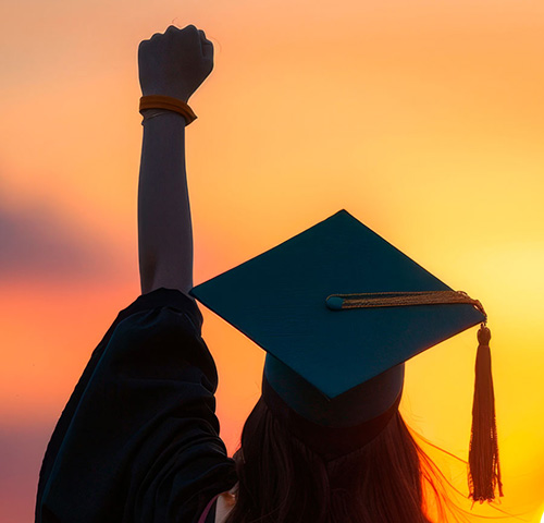 KSU Graduation celebration at sunset with a female student in cap and gown raising her fist in triumph against a vibrant orange sky.