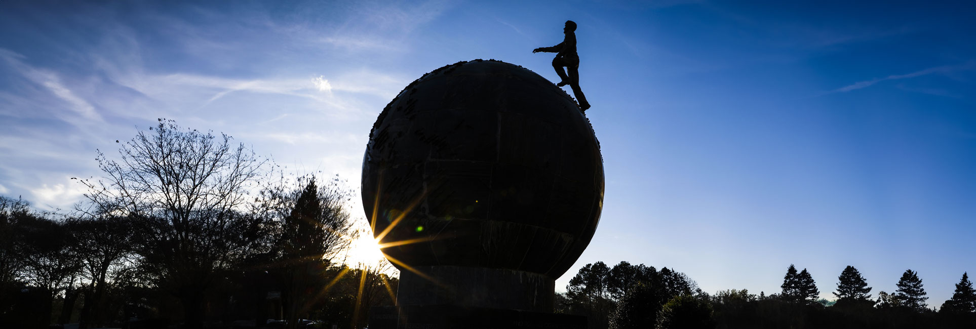 Silhouette of a person walking on top of the world globe sculpture on the Kennesaw State campus with a sun setting behind it. Trees and a clear blue sky are visible in the background.