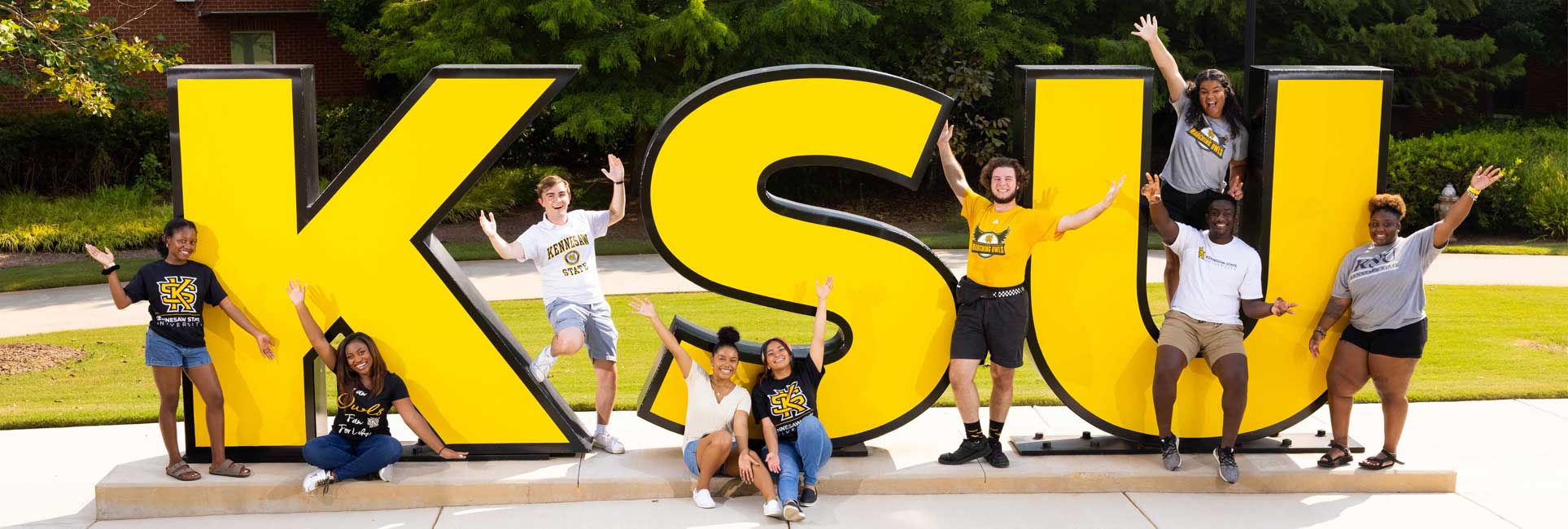 A group of diverse Kennesaw State University students posing around and on top of large, bold yellow letters spelling "KSU" outside on campus. The students are smiling and holding their arms up in excitement, wearing various KSU-branded clothing. The scene is bright and energetic, set against a green landscape with trees in the background.