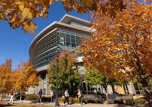 A modern, curved glass building on the Kennesaw State University campus surrounded by vibrant autumn trees with golden and orange leaves. Students walk along a path in front of the building, enjoying the crisp fall day under a bright blue sky. The scene captures the beauty of the campus during the fall season.