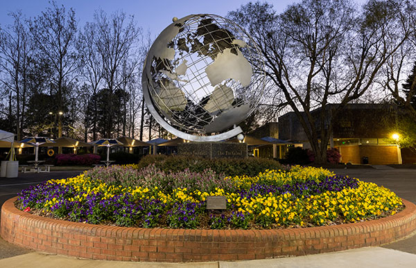 A large metal globe sculpture on the Kennesaw State University Marietta campus, set in the middle of a circular brick flower bed filled with vibrant yellow and purple flowers. The globe is surrounded by a well-maintained walkway, with trees and campus buildings visible in the background during dusk, adding a peaceful ambiance to the scene.