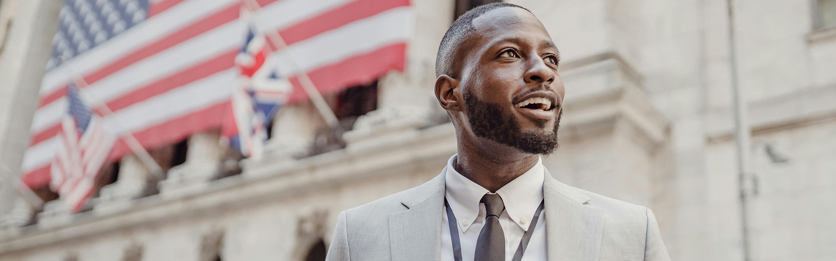 Professionally dressed black man stands in front of government building with flags in the background.