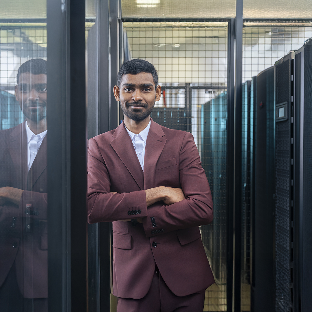 A man in a suit stands confidently in front of a server rack filled with equipment.