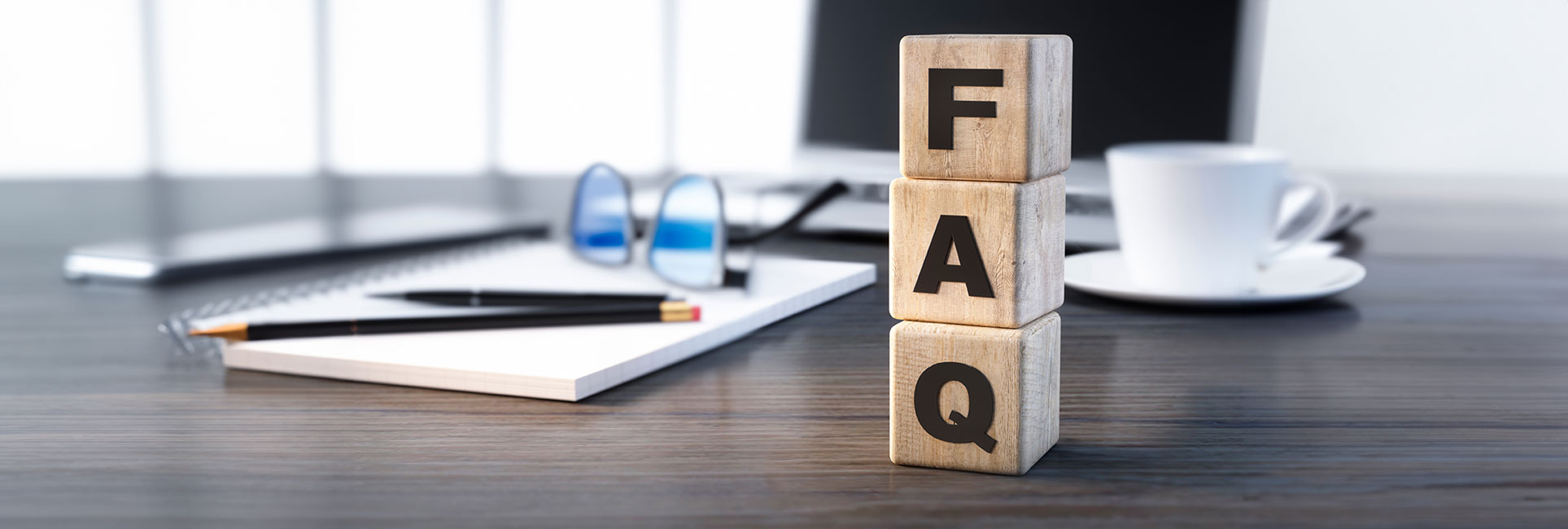 pelling out "FAQ." The blocks are on a wooden desk with a notebook, pen, glasses, and a coffee cup in the background. A laptop is partially visible on the desk.