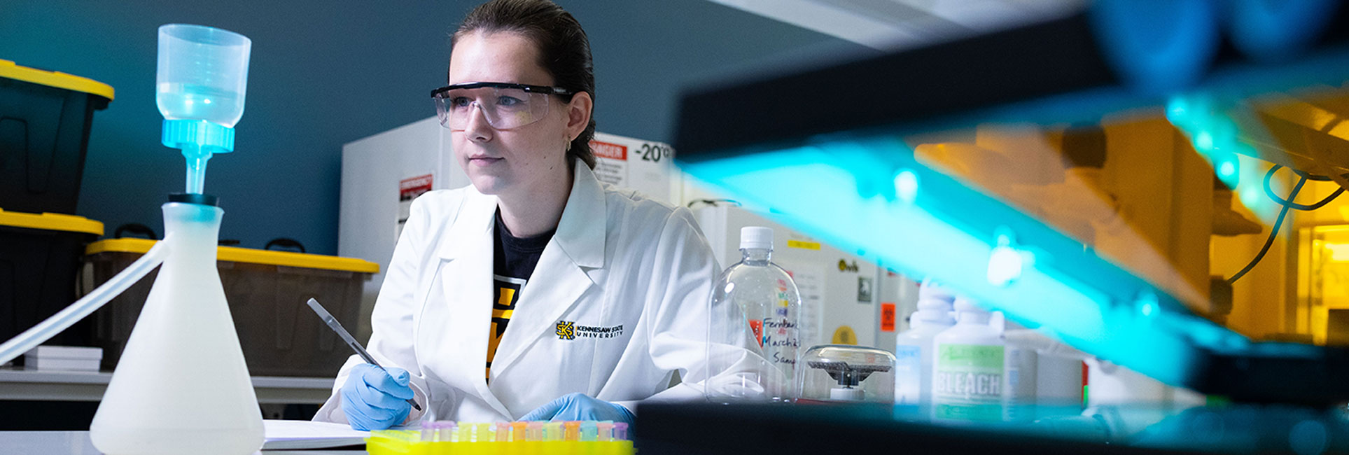 A KSU graduate student wearing safety goggles and a Kennesaw State University lab coat works diligently in a laboratory. She is using a pipette while analyzing samples, with various lab equipment, such as a filtration flask and a UV light, surrounding her. The setting emphasizes precision and focus in scientific research.