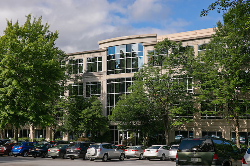 Kennesaw State Town Point building is a modern, three-story office building with a curved glass facade and a flat roof. The building is surrounded by green trees and a parking lot filled with cars. The sky is blue with some white clouds.