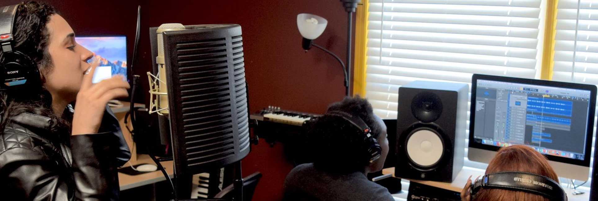 A recording studio setting featuring a woman wearing headphones and singing into a professional microphone with a pop filter. In the background, two individuals wearing headphones are seated at a workstation, working on a computer displaying audio editing software.