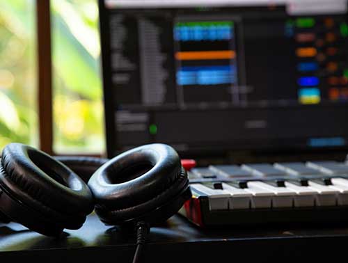 Close-up of a music production setup, featuring a pair of black headphones resting on a keyboard controller. In the background, a laptop screen displays music editing software with colorful tracks and menus. 