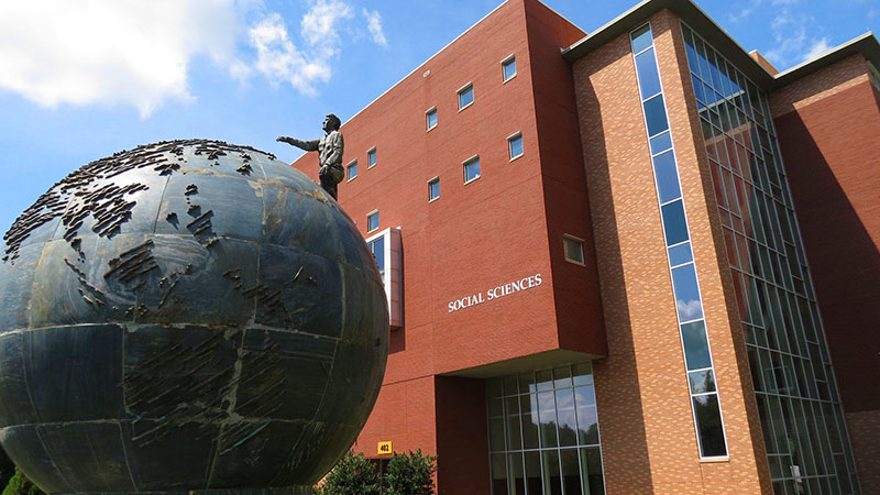 A large globe sculpture with a statue of a man standing on top of it, in front of a red brick building labeled "Social Sciences."