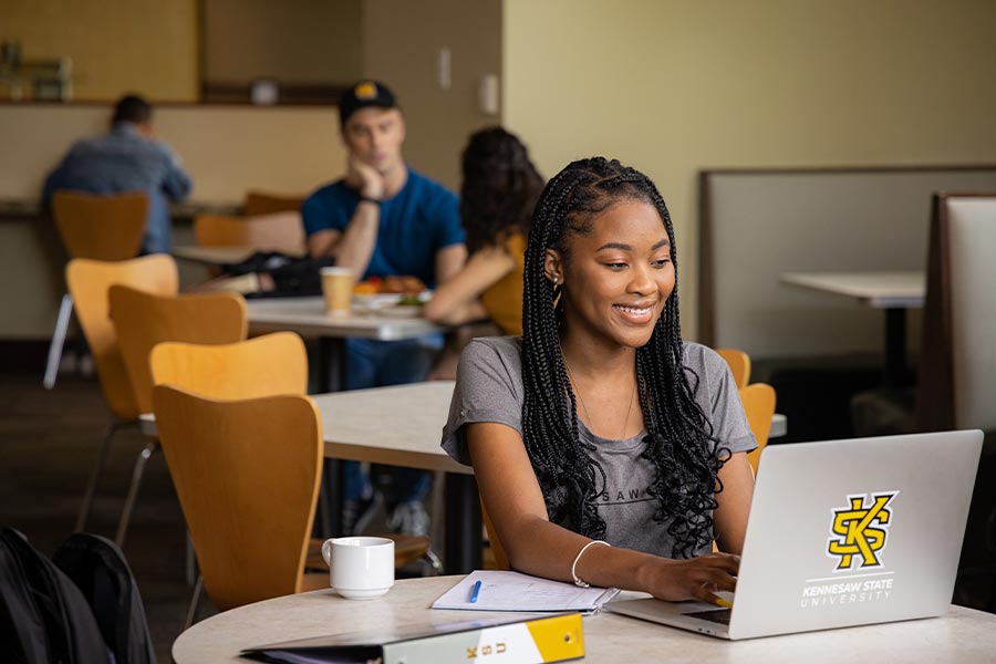 ksu student in a public area at a table using their laptop.