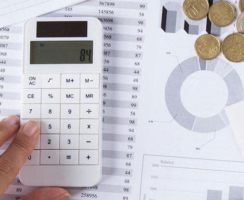 A calculator with a hand hovering over the buttons, placed on top of financial documents. The documents include graphs, charts, and numbers, along with a group of coins.