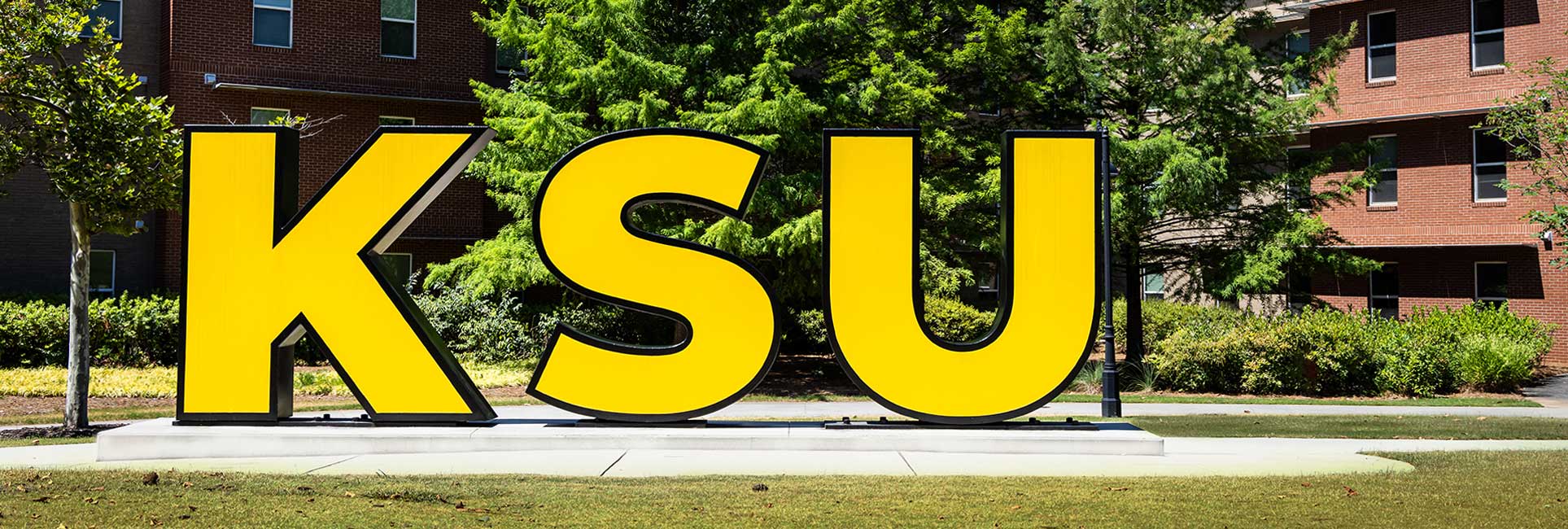 Large, gold letters spelling out "KSU" (Kennesaw Sate University)in front of a campus building and green trees.