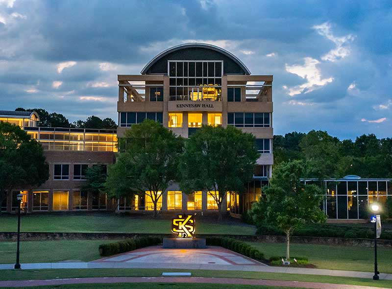 Kennesaw Hall, at Kennesaw State University is a modern brick building with a large glass dome, stands illuminated against a twilight sky. The building is surrounded by green lawns and trees.