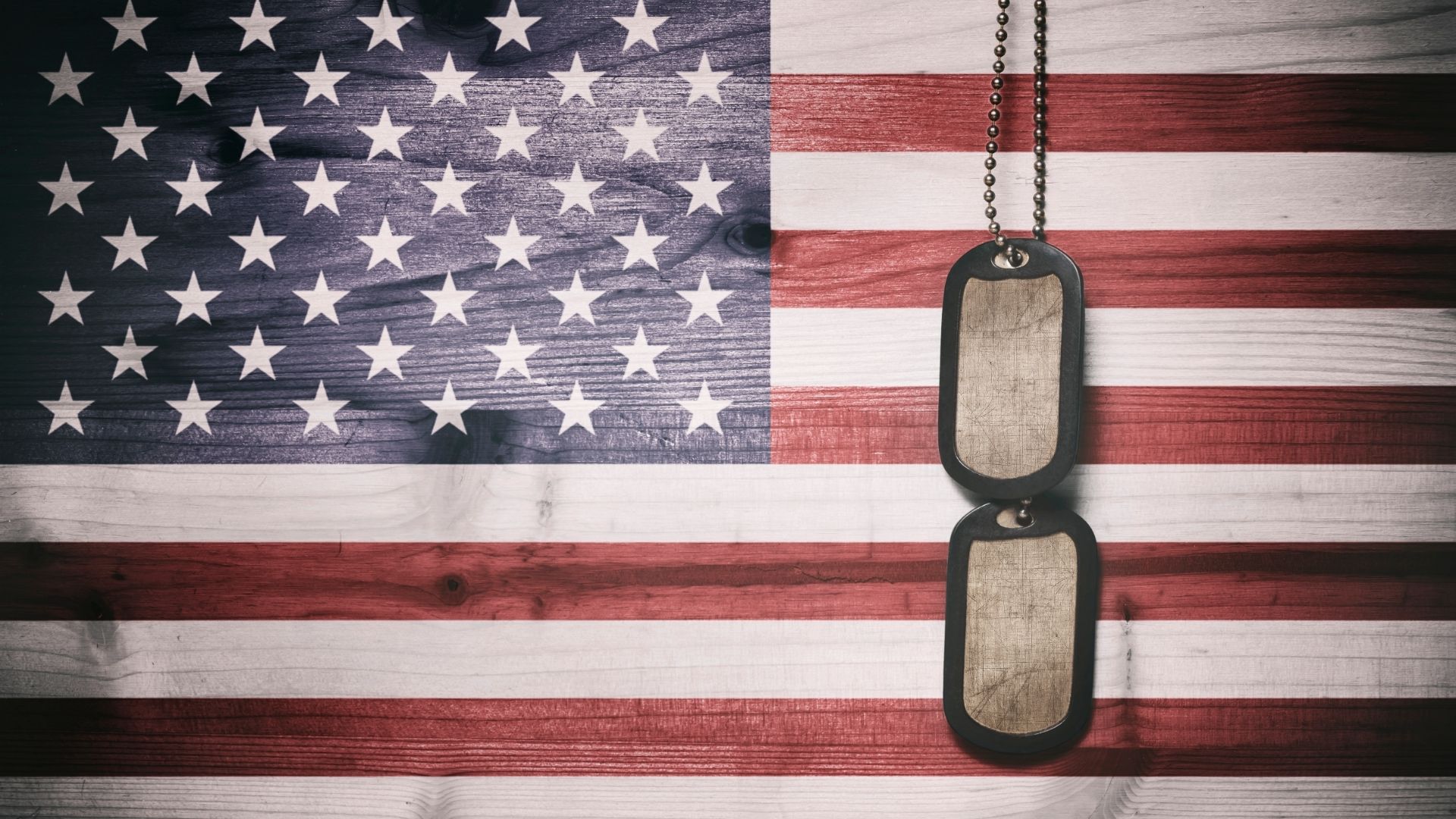 Two military dog tags hang from a chain in front of an American flag on a wooden background.