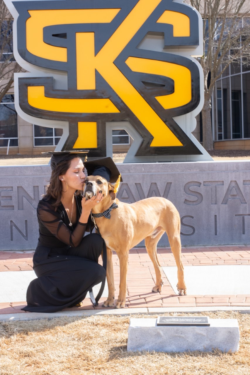 mary jones kissing her dog in front of the KSU Logo sign