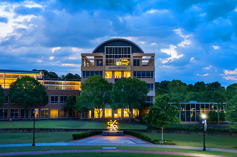 Kennesaw Hall stands out against a twilight sky. The building has a modern design with a large, curved glass facade and a smaller, illuminated dome on top. Trees frame the building on either side, and a grassy lawn stretches out in front.