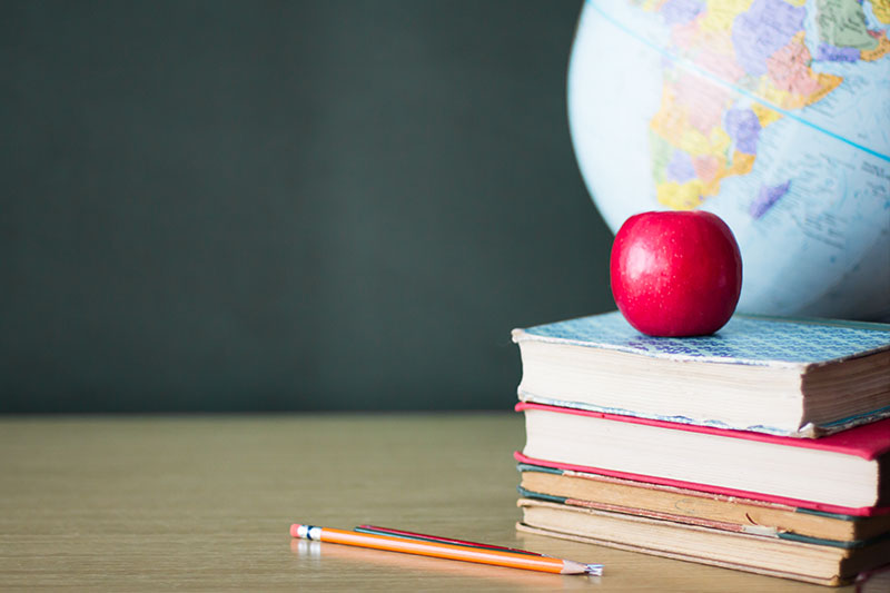 Stacked academic books on a table with an apple on top of the books and a world globe in the background