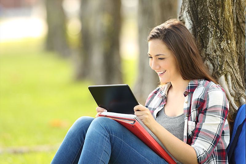 Female student watch videos on a tablet outside sitting at a tree