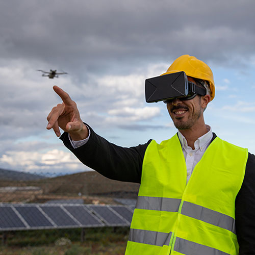 A civil engineer wearing a virtual reality headset and a yellow safety vest, pointing towards a flying drone, with solar panels and cloudy skies in the background.