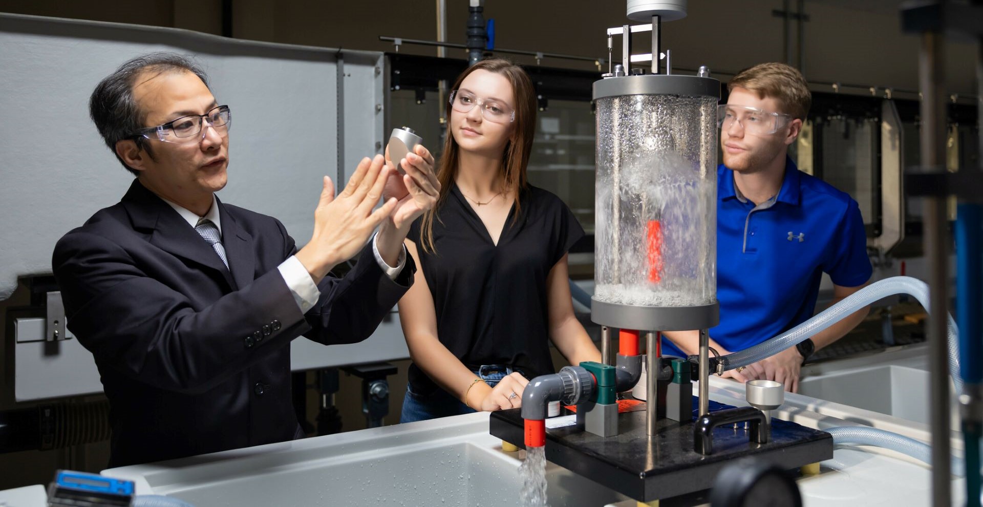 An instructor at KSU demonstrates a device to two students wearing safety goggles, standing next to a fluid dynamics experiment in a laboratory.