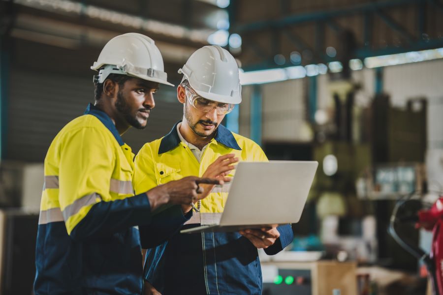 Two engineers in safety gear collaborating while using a laptop in an industrial setting.