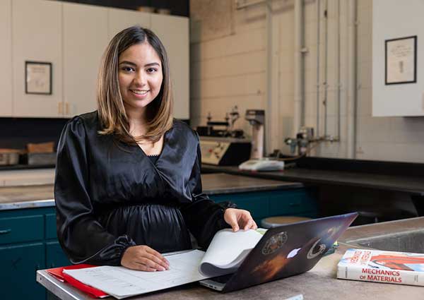 KSU engineer graduate, Maria Gonzalez sitting at a desk with a laptop and holding a piece of paper.