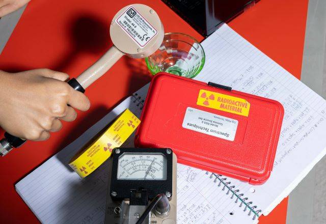 Hand using a radiation detection device near a red case labeled 'Radioactive Material,' surrounded by notes and measurement tools.