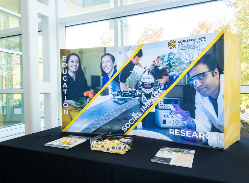 KSU SPCEET scholarship reception area with a table and a poster sitting on top of the table, displaying the words, "education, social impact, and research." 